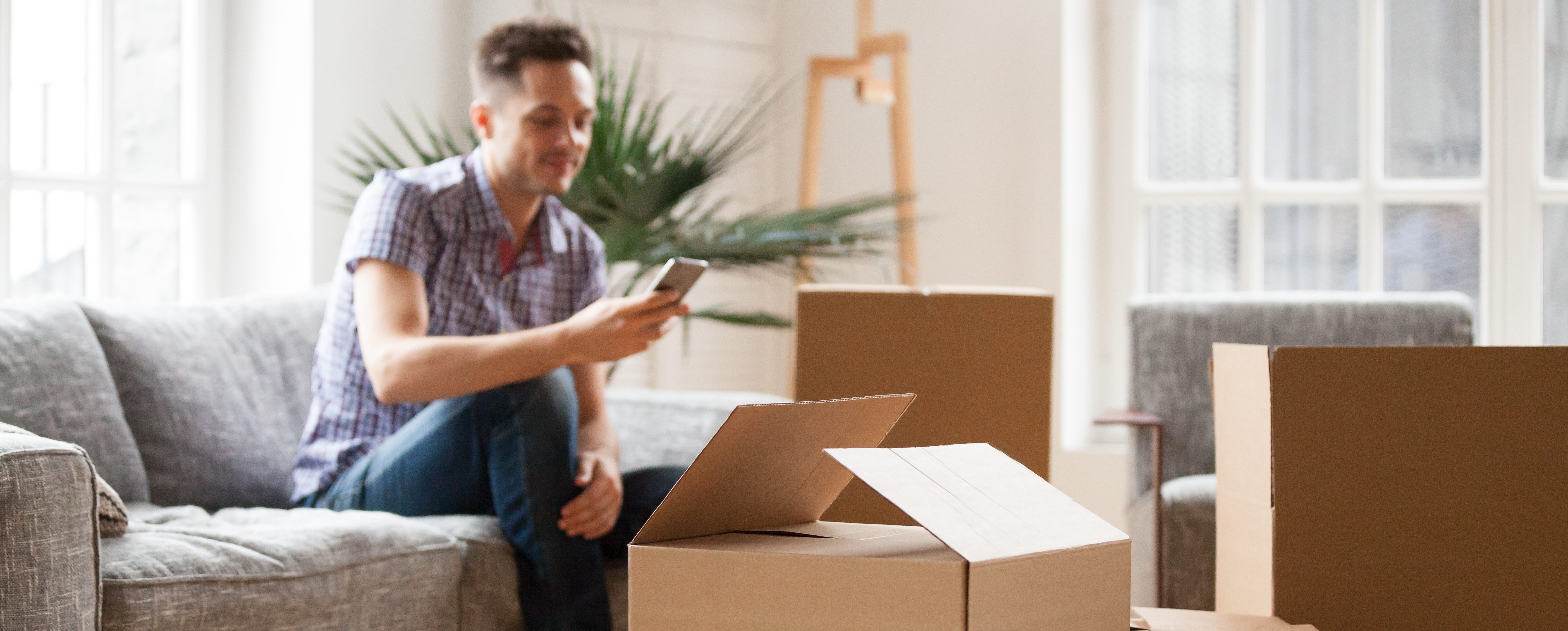Packed cardboard boxes with young man sitting on sofa in living room calling delivery service at background, belongings in carton containers waiting for moving in out new home or relocation concept